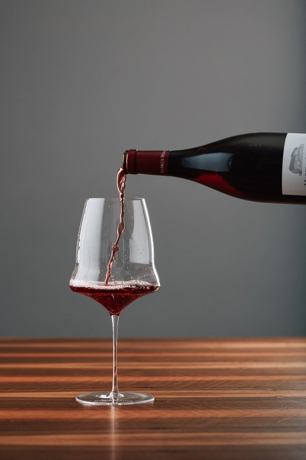 Roter Wein wird aus einer Flasche in ein Glas auf einem Holztisch gegossen vor grauem Hintergrund